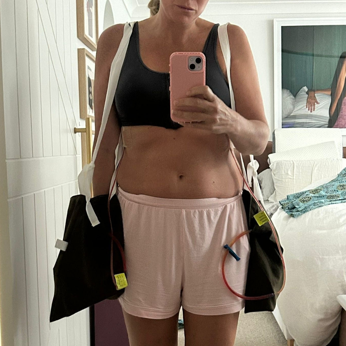 A woman takes a mirror selfie in a bedroom setting, wearing a black sports bra and light pink athletic shorts, with black duffel bags slung over her shoulders.